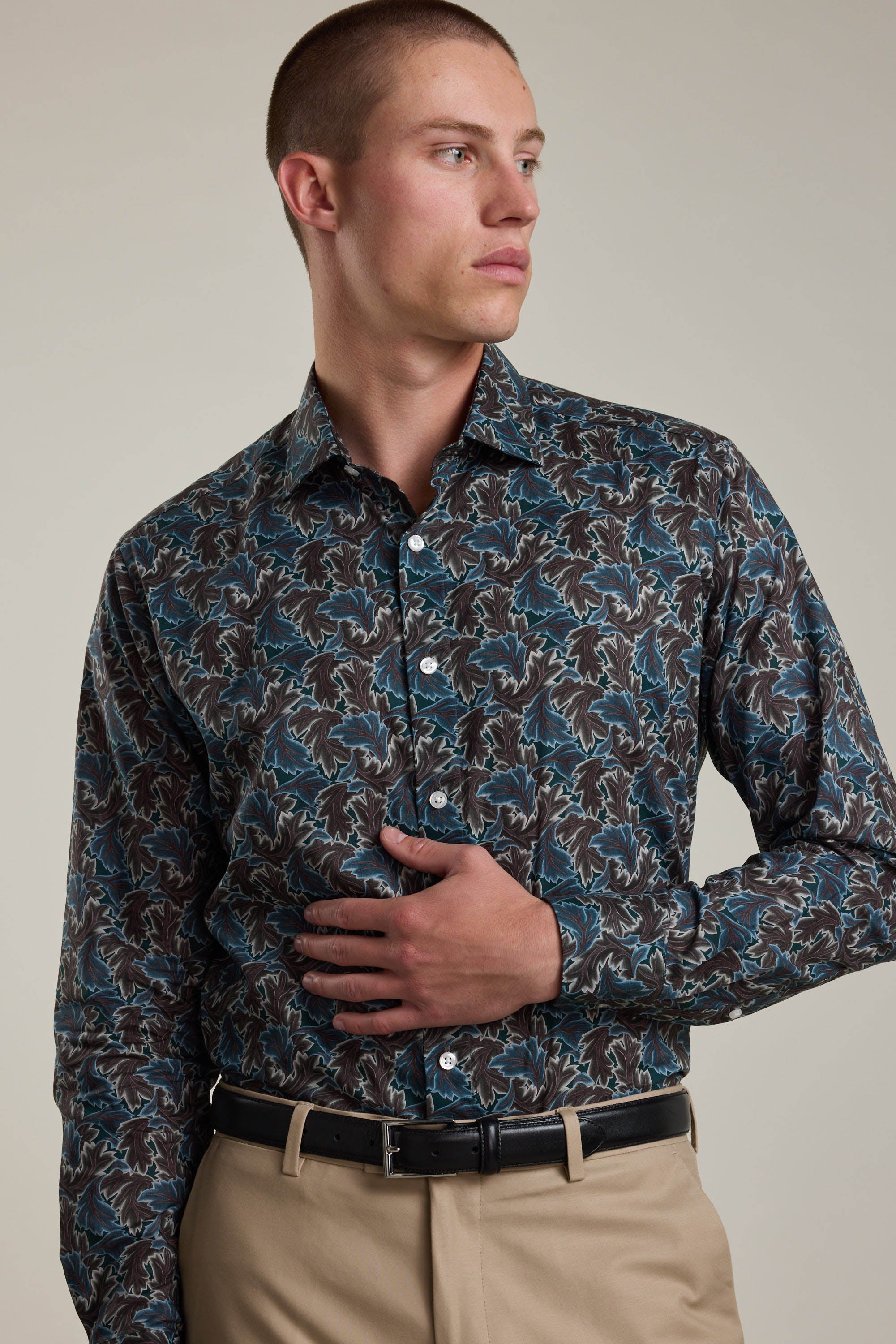 A man in a Liberty Midnight Canopy Shirt by Liberty—a patterned long-sleeve in blue, brown, and black—paired with beige pants and a black belt, stands with one hand on his stomach, looking to his left.