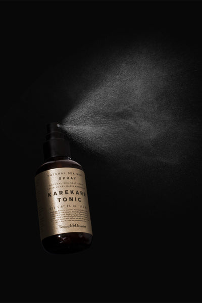 A dark amber spray bottle of Triumph & Disaster Karekare Tonic Salt Spray releases a fine mist against a black background. The label highlights it as a natural sea salt spray by Triumph & Disaster for effortlessly textured hair.