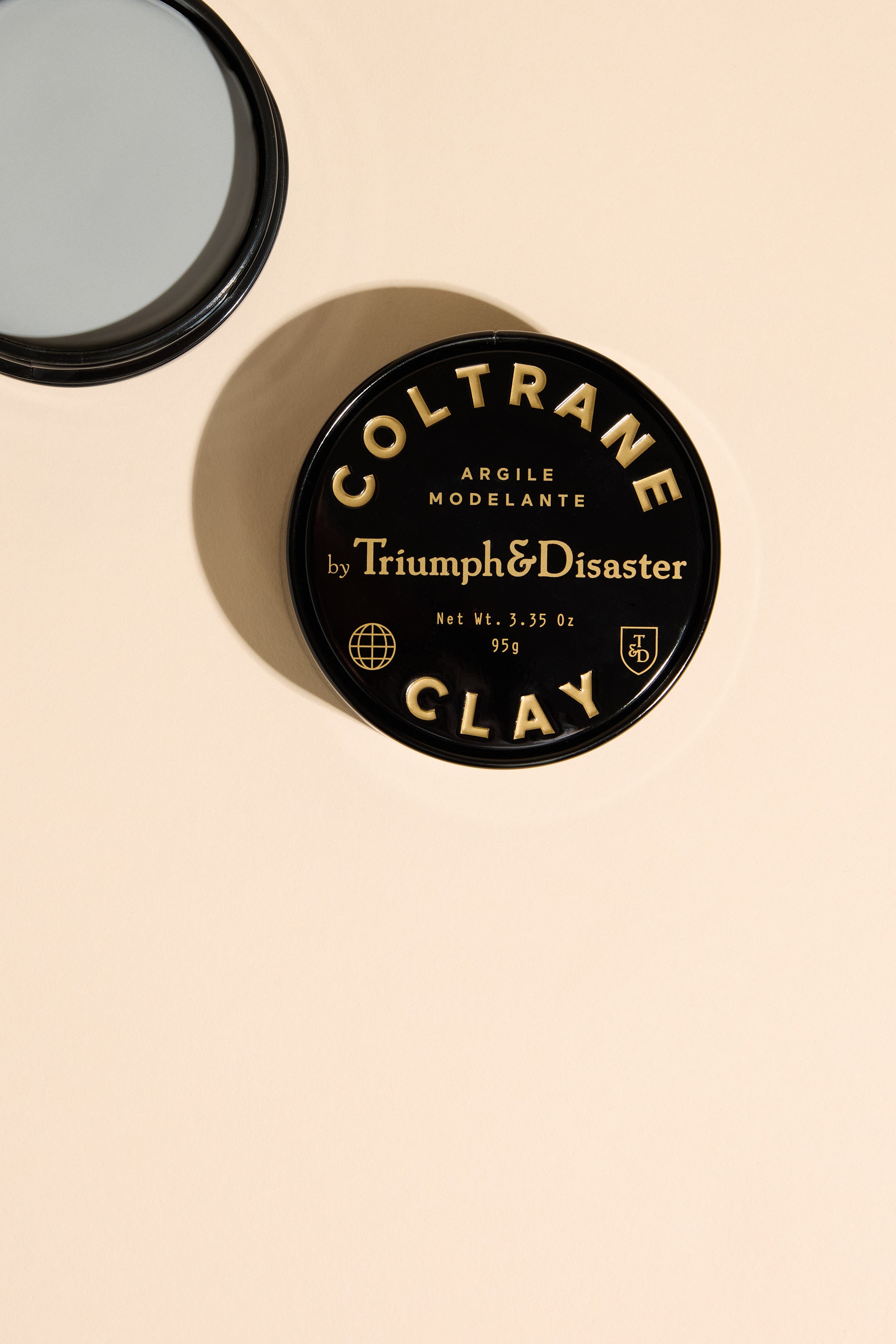 A round black tin of Triumph & Disaster Coltrane Clay, a matte hair styling clay, sits on a beige surface beside a small mirror partially visible in the top left corner.