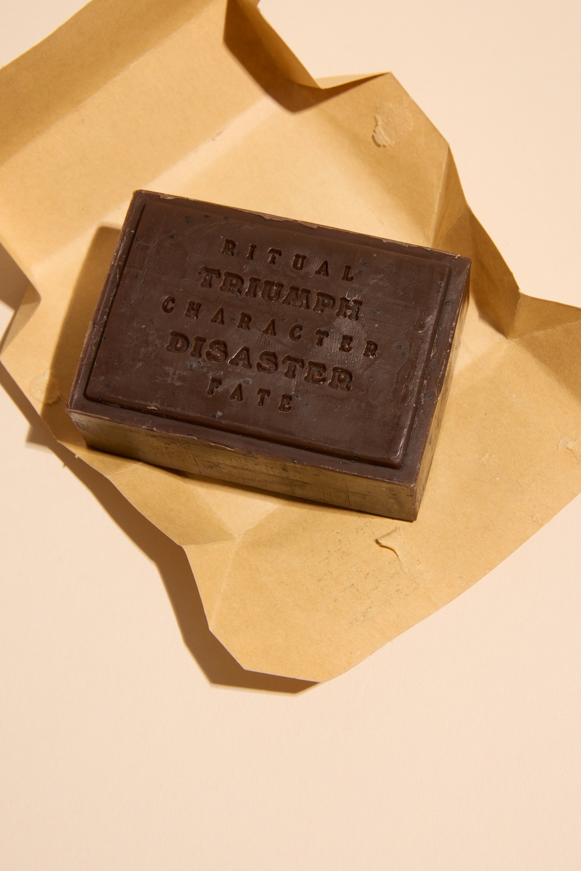 A rectangular dark chocolate bar sits on brown parchment, embossed with words like RITUAL, TRIUMPH, and FATE—inviting a moment as comforting as Triumph & Disaster Shearers Soap, valued for its gentle touch.