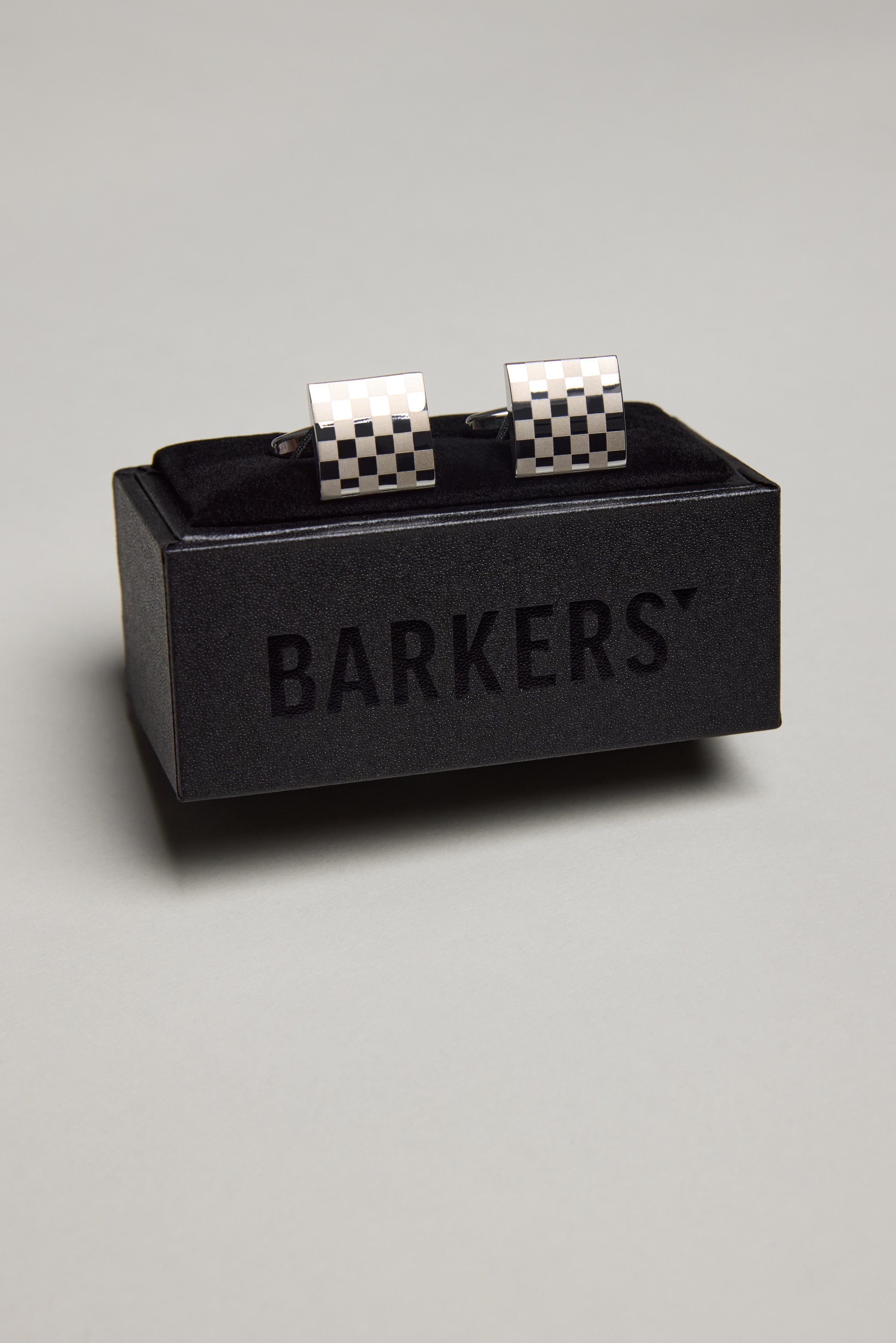 The Classic Silver Chequered Cufflink by Barkers features a polished finish and checkered pattern, displayed on a black velvet cushion in a black Barkers box against a plain light grey background.