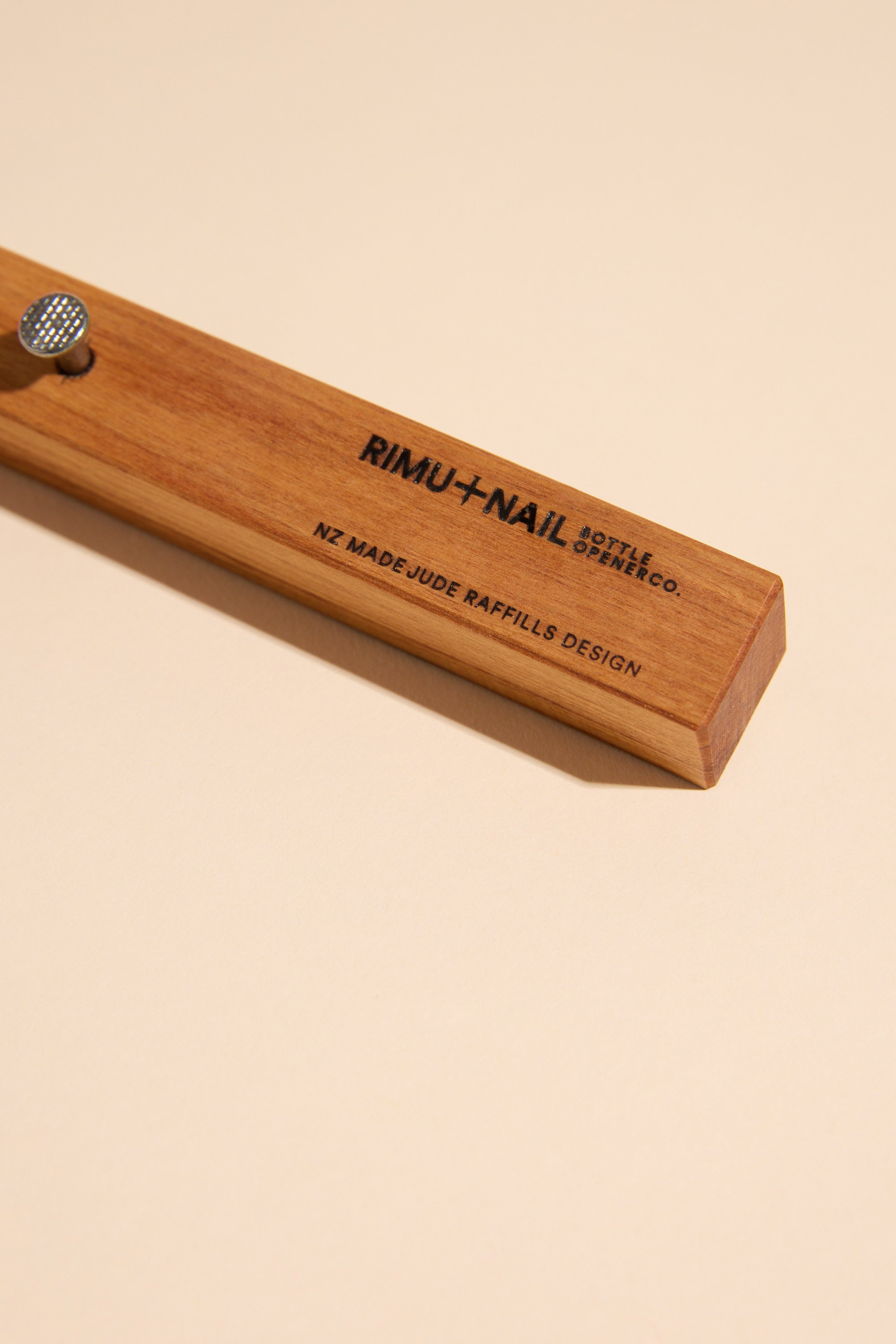 The Rimu & Nail Bottle Opener by Other, handmade in NZ from rimu and stamped with black branding, lies on a beige surface.