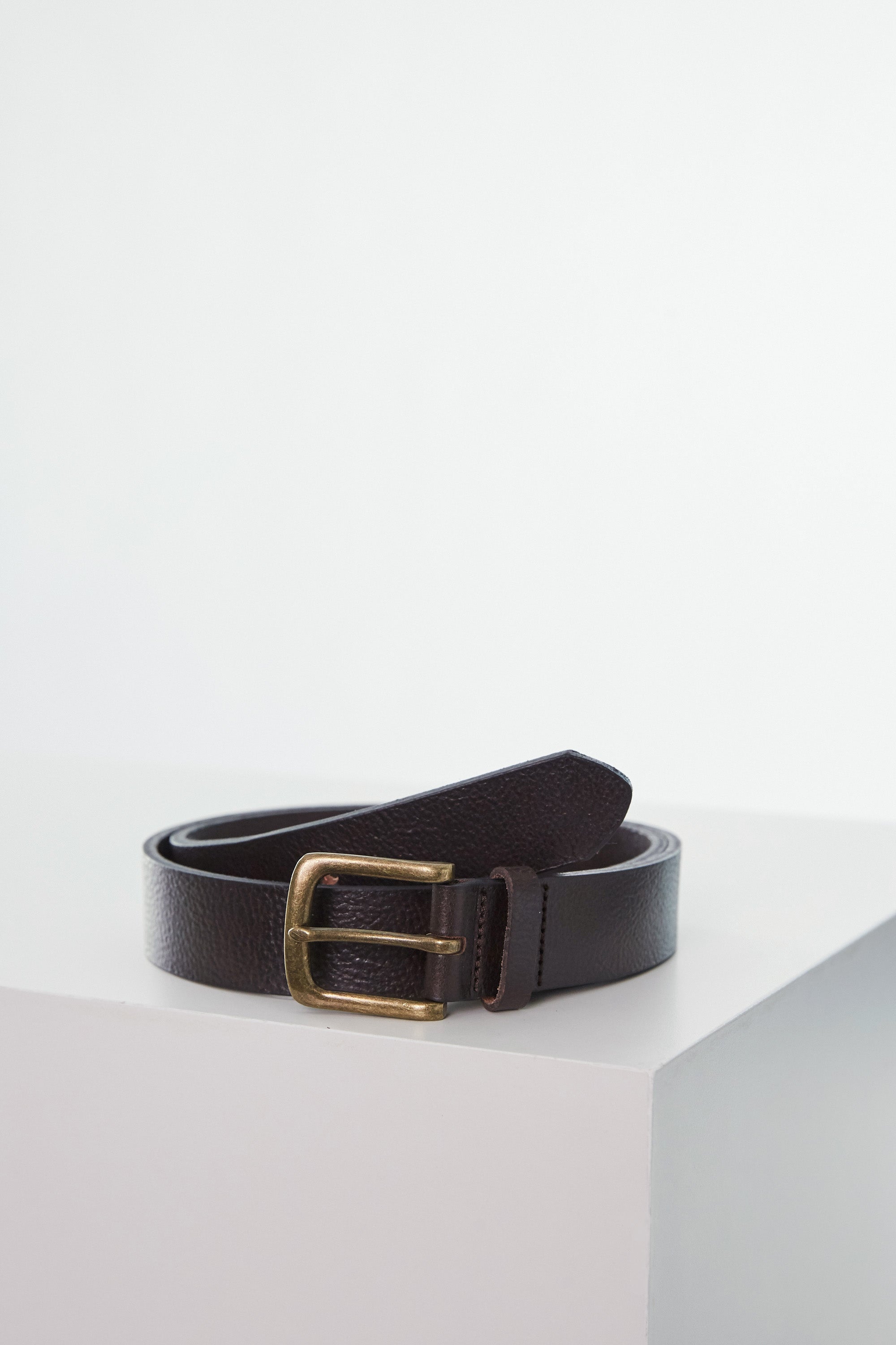The Baywood Tumbled Leather Belt by Barkers, featuring a goldtone buckle and rich dark brown leather, is coiled neatly on a white surface, its color standing out against the plain background.