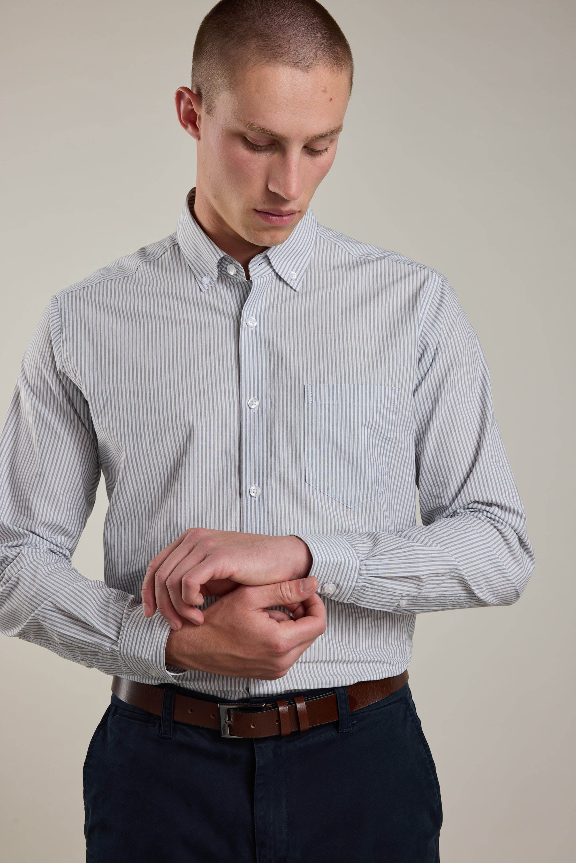 A man in the Barkers Miller Stripe Shirt, a light blue cotton poplin tailored-fit style, and dark pants adjusts his sleeve cuff while looking down against a plain, light background.