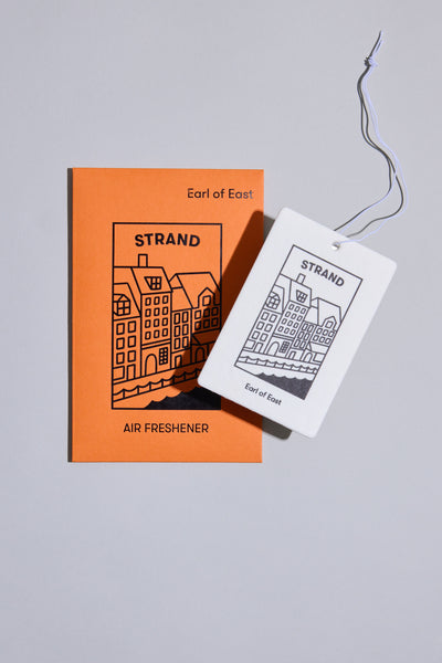 The EoE Strand Air Freshener by Earl of East features a line drawing of buildings on both the rectangular scented freshener and its orange card, with branding from Earl of East and STRAND for a stylish, portable fragrance.