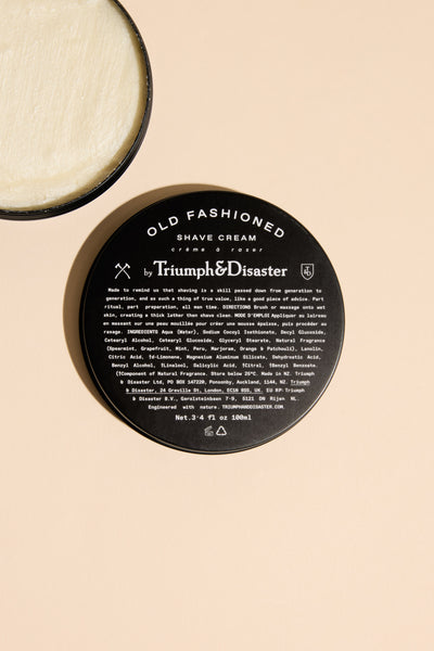A round, black jar of Triumph & Disaster Old Fashioned Shave Cream is shown open on a light beige background, displaying its rich cream—an essential from the Triumph & Disaster grooming range.