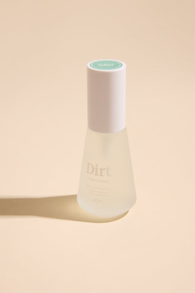 A clear frosted glass Carry On Fabric Spray by Other, with a white cap and green label, sits on a beige surface casting a soft shadow. This 30ml spray uses probiotic technology to eliminate odours from fabrics.