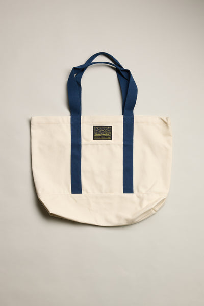 The Barkers Supply Co. Canvas Tote Bag by Barkers is a durable cream canvas tote with two blue handles, matching vertical stripes, and a small rectangular logo patch—perfect for everyday travel use.