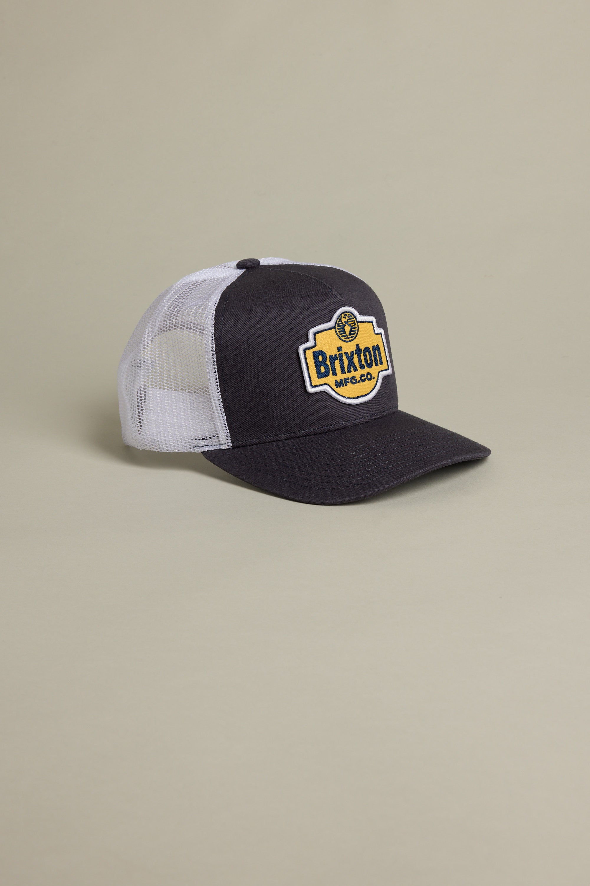 The Brixton International Trucker by Brixton is an eco-friendly black and white mesh trucker hat with a yellow and black patch on the front, shown against a neutral beige background.