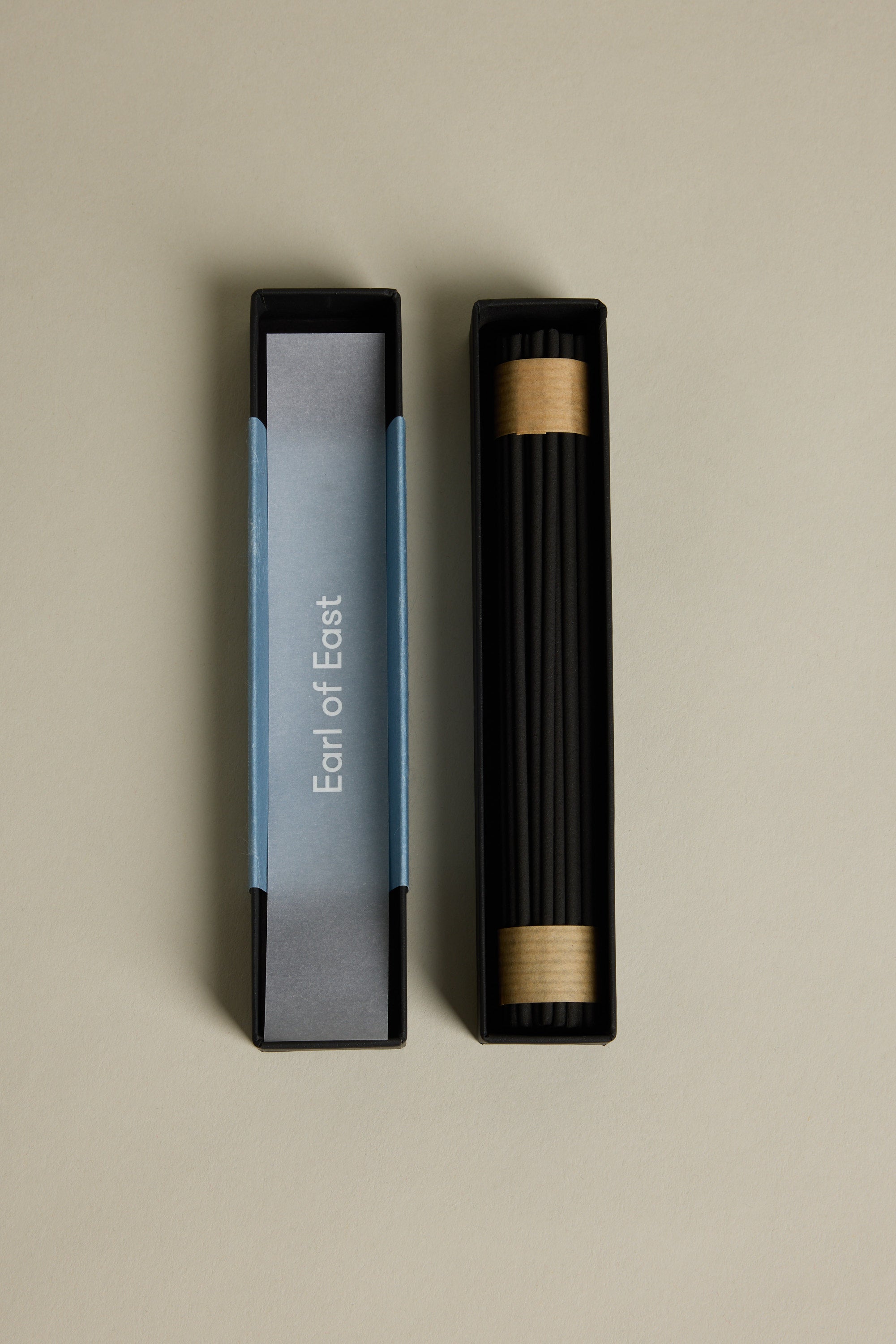 A rectangular black box labeled “Earl of East” holds EoE Onsen Japanese Incense Sticks, each wrapped at the ends with brown paper. The open box and lid rest on a beige surface, inviting a calming Japanese-inspired ritual.