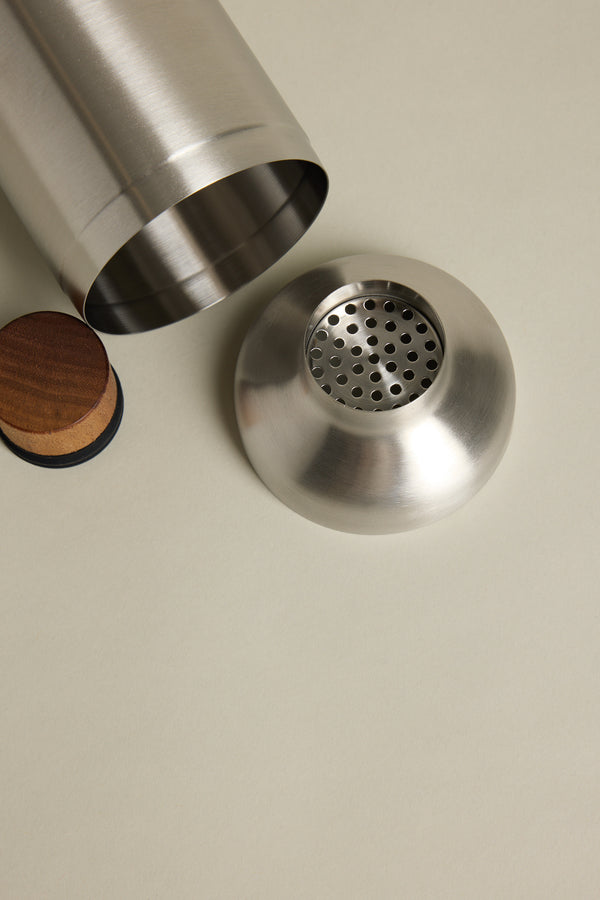 The Other Liquid Lounge Cocktail Shaker, with its lid and strainer removed, rests on a light beige surface beside a round wooden coaster.