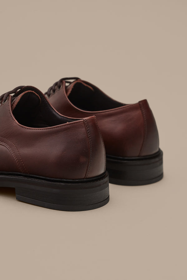 A close-up of the back and side of Barkers' Saville Derby Shoe, a brown leather men’s dress shoe with black soles, set against a beige background, highlighting quality Portuguese craftsmanship.