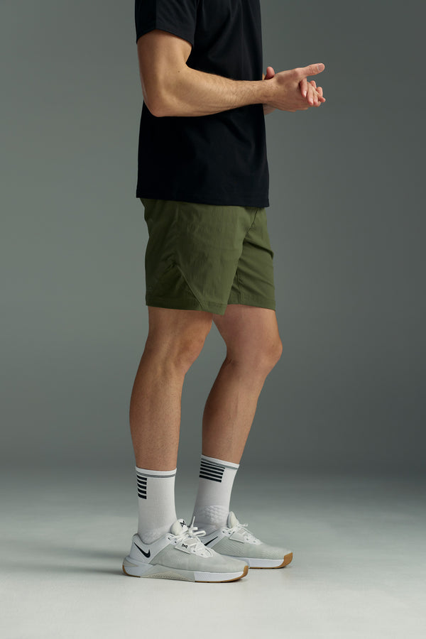 A person stands indoors wearing a black shirt, the WearEver Agile Short by WearEver (lightweight with 7-inch inseam), white athletic socks with black stripes, and white sneakers. Their hands are loosely clasped and their face is not visible.