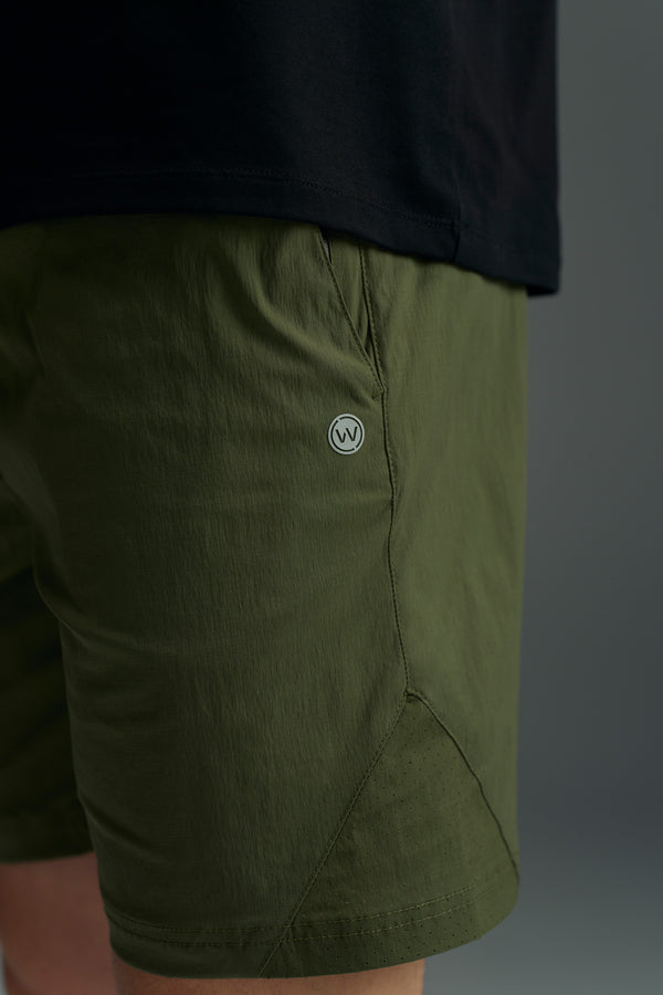 A close-up of a person wearing WearEver Agile Shorts in olive green, featuring a small circular logo near the pocket, paired with a black shirt against a plain gray background.