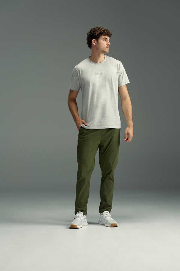 A man stands with one hand in his pocket, wearing the WearEver Outbound T-Shirt by WearEver in light gray, olive green pants, and white sneakers. He poses against a plain gray background, looking off to the side with a relaxed posture.