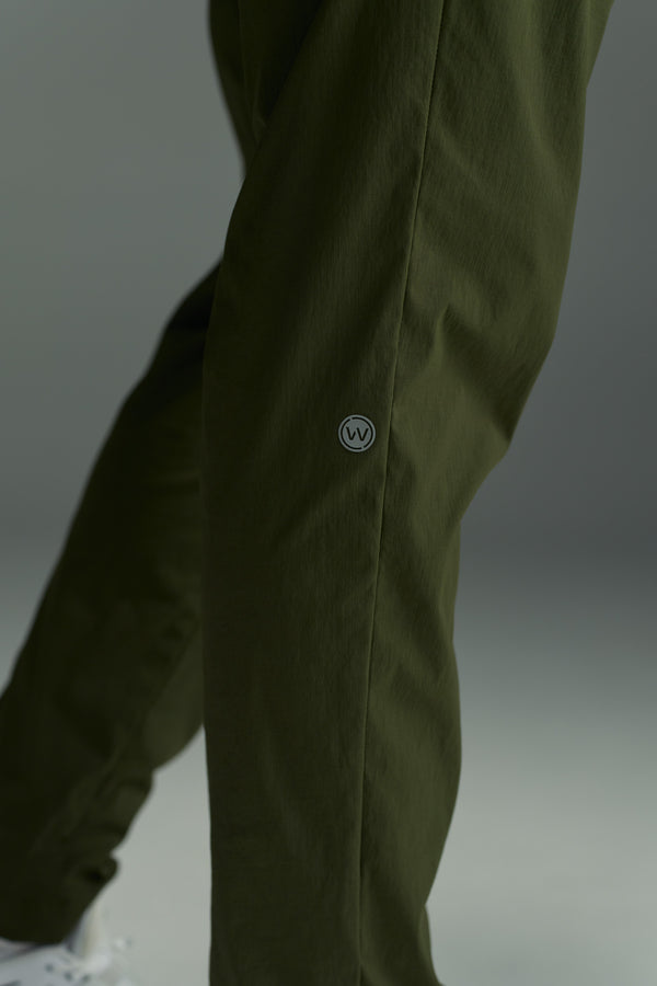 A close-up of someone wearing WearEver's olive green Transit Pant, featuring a small circular logo on the thigh and part of a white shoe visible in the lower left.