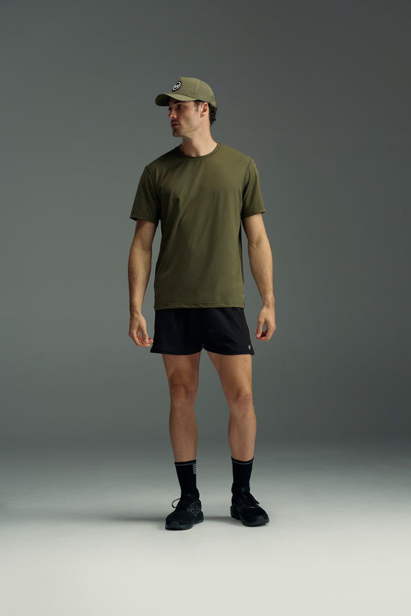 A man wearing the WearEver Pulse Tee in olive green, with black athletic shorts, socks, and sneakers stands against a plain gray background, looking to the left.