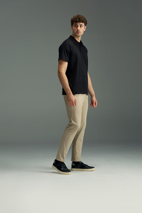 A man stands on a plain background wearing a black polo shirt and WearEver Long Game Pant by WearEver, made with lightweight, quick-dry fabric. He looks slightly to the side with one arm relaxed and the other slightly bent.