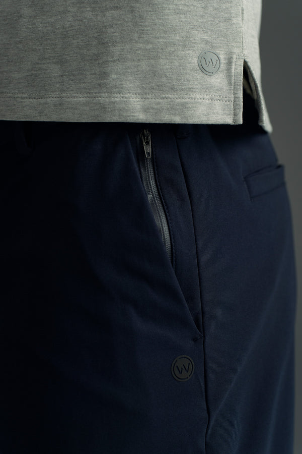 Close-up of a person in a light grey shirt and WearEver Long Game Pant by WearEver in dark navy with a visible zipper pocket. Both feature subtle circular logos, highlighting the chino-inspired style and performance fabric texture.
