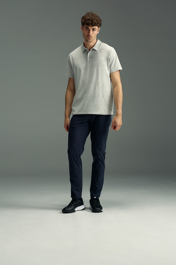 A man with curly hair wears a light gray polo, black sneakers, and dark navy WearEver Long Game Pant by WearEver, standing against a plain gray background.