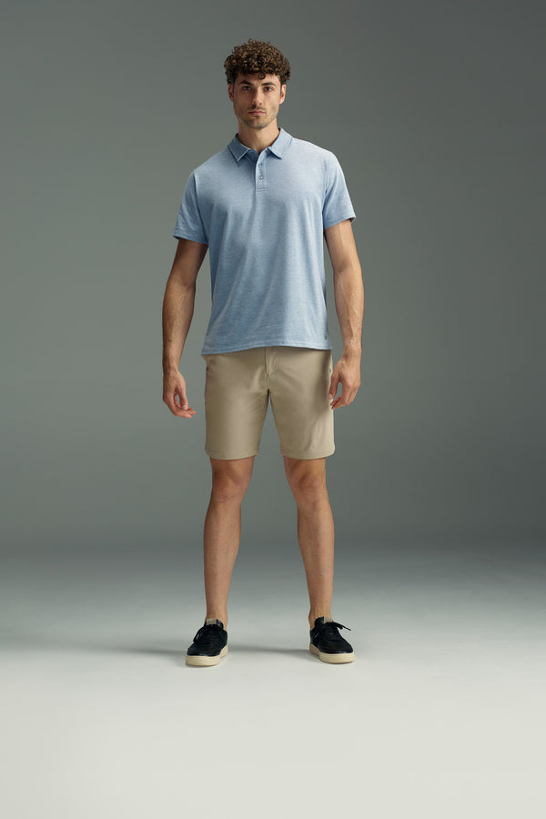 A man stands facing forward, wearing a light blue polo, black shoes, and beige WearEver Switch Shorts by WearEver, against a plain gray background.