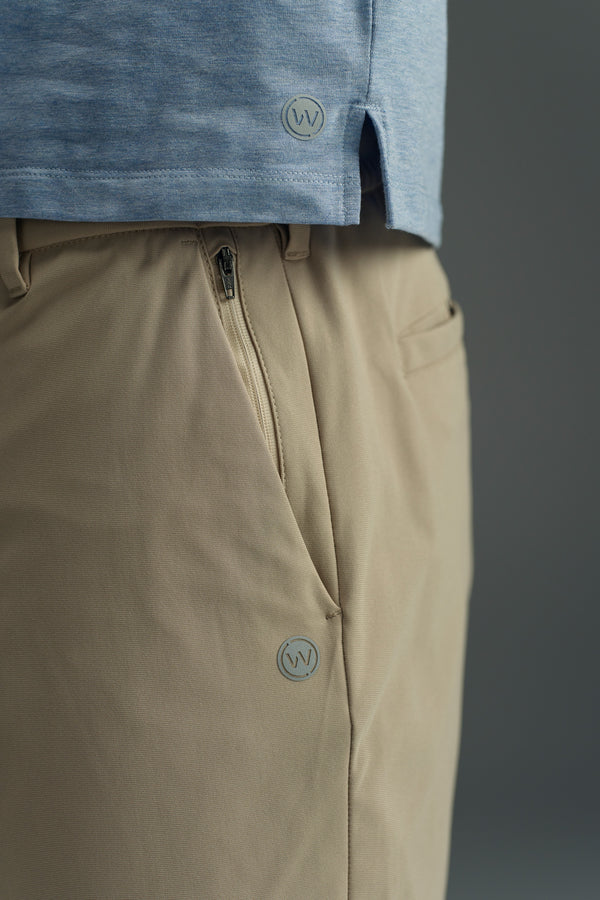 Close-up of a person wearing a light blue shirt and WearEver Switch Short by WearEver in beige with comfort stretch, featuring a visible zipper and small circular logos. The plain background is out of focus.