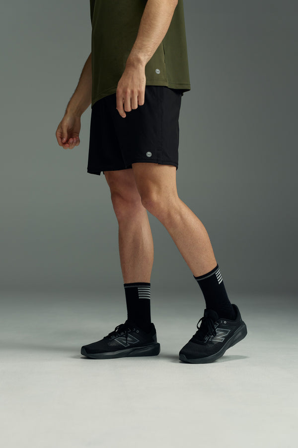 A person stands in a studio wearing a green shirt and black WearEver Cadence Short with a 5-inch inseam, paired with black socks featuring white stripes and black running shoes. The upper body is cropped out of the frame.