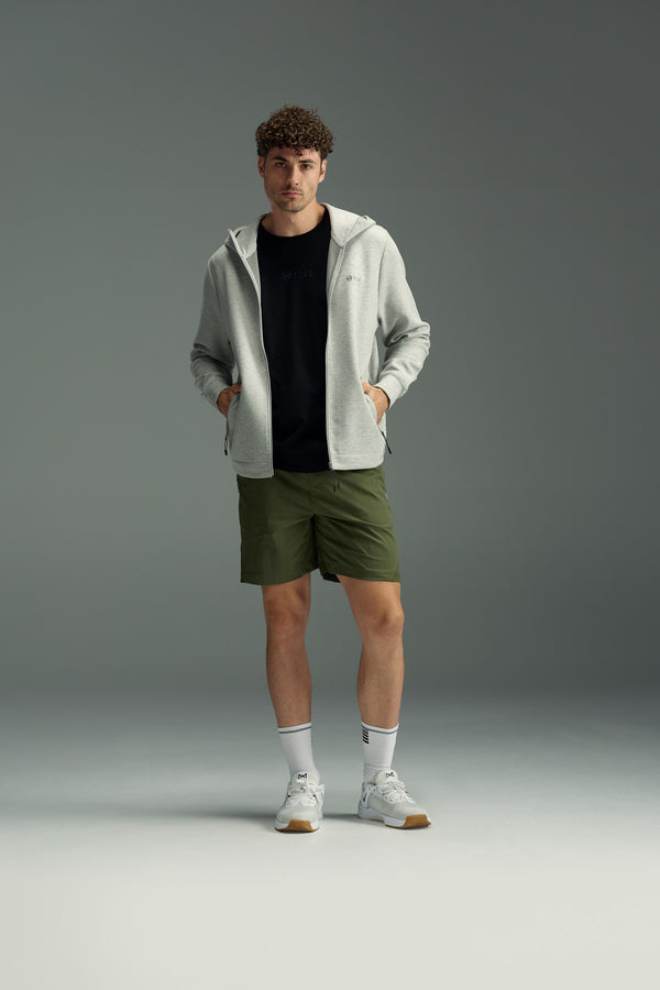 A man stands against a neutral background wearing the WearEver Altitude Hoody from WearEver, paired with a black t-shirt, olive green shorts, white crew socks, and white athletic sneakers. His hands rest in his hoody’s pockets.