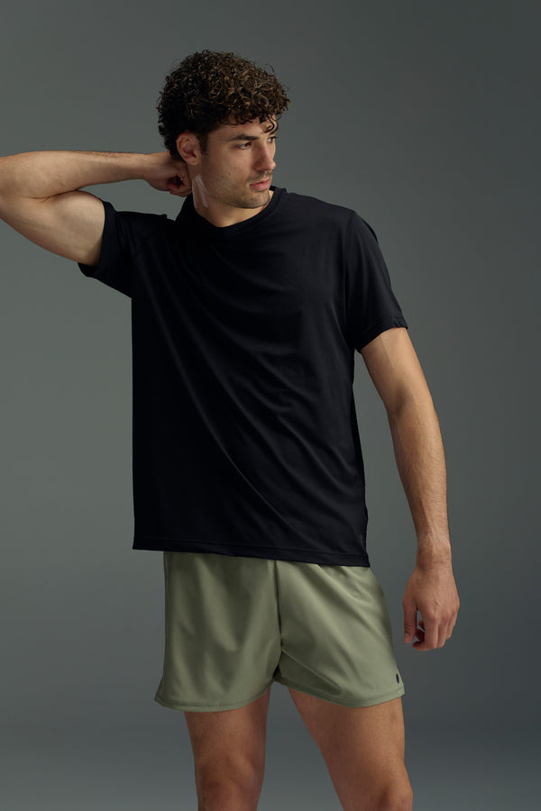 A man with curly hair stands against a plain background, wearing the WearEver Pulse Tee in black and light olive green athletic shorts. He has one arm bent behind his head and looks to the side.