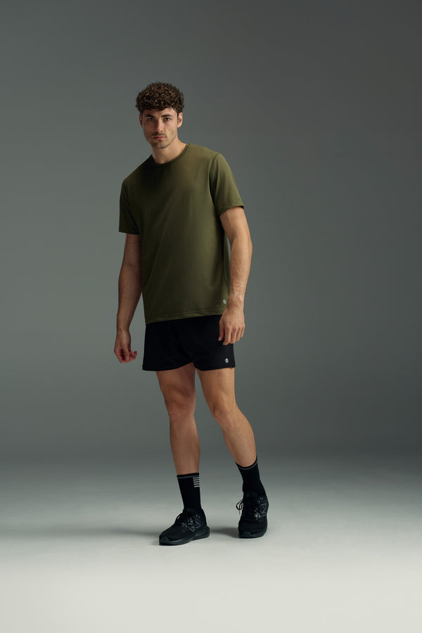 A man stands in a studio wearing the olive green WearEver Pulse Tee by WearEver, paired with black shorts, socks, and sneakers. He faces forward with a neutral expression against a plain gray background.