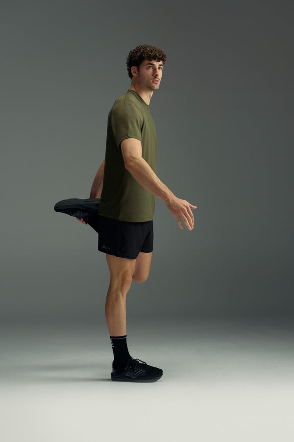 A man wears the WearEver Pulse Tee in olive green and black shorts as he performs a quad stretch, standing on one leg and looking upward in a neutral studio setting.