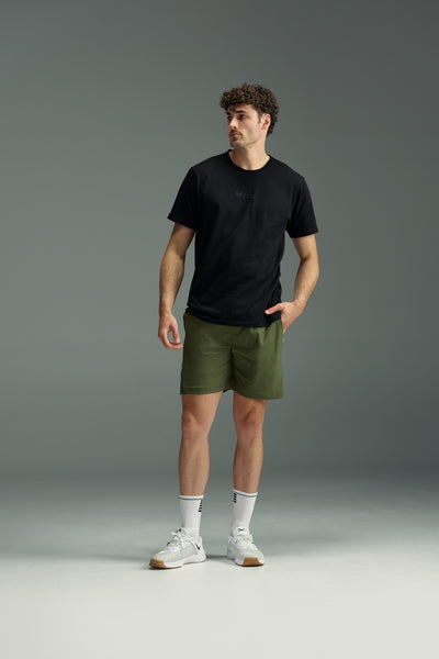 A man stands against a plain gray background wearing a black t-shirt and olive green WearEver Agile Short by WearEver, featuring lightweight stretch fabric and a 7-inch inseam. He pairs them with white crew socks, white sneakers, and has one hand in his pocket.