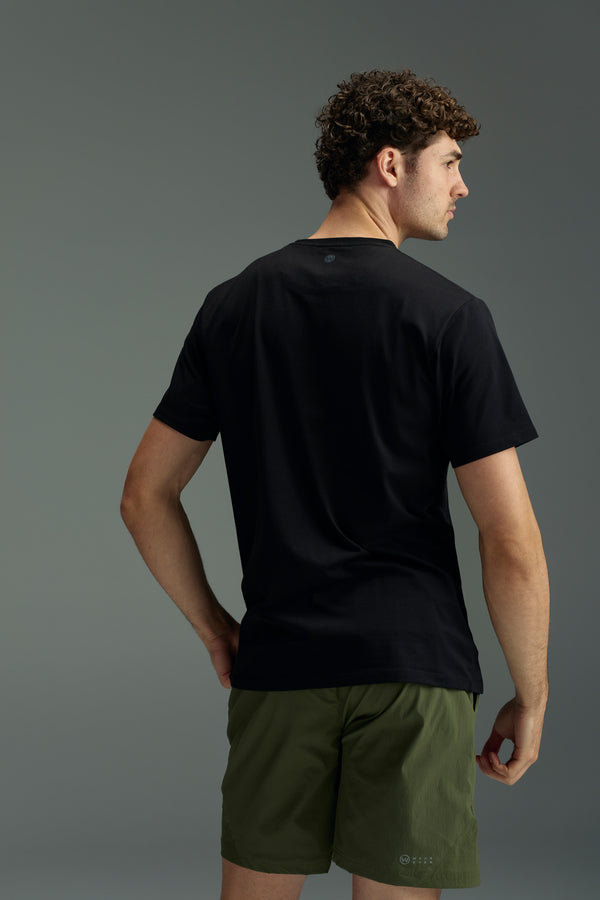 A man with curly hair faces away, wearing the WearEver Outbound T-Shirt by WearEver and olive green shorts against a plain gray background. His left hand is on his hip.