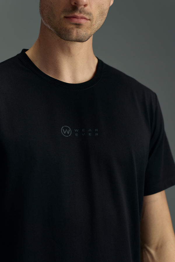 A man wears the WearEver Outbound T-Shirt by WearEver, a black cotton stretch tee featuring a subtle chest logo, photographed against a plain grey background with his face partially visible.