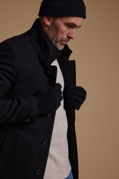 A man in a black coat and gloves stands against a beige background, adjusting his coat. He wears a Woolyarns Perino NZ Made Perino Beanie, has a trimmed beard, and looks down thoughtfully.