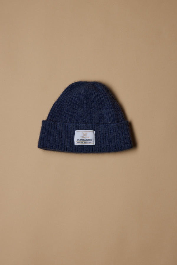 The NZ Made Perino Beanie by Woolyarns Perino is a dark blue knitted merino possum silk blend beanie with a folded brim and white front label, shown against a plain beige background.