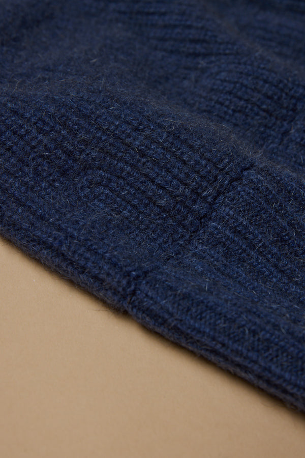 Close-up of the NZ Made Perino Beanie by Woolyarns Perino, featuring dark blue ribbed knit fabric in a luxurious merino possum silk blend, shown on a light brown surface.