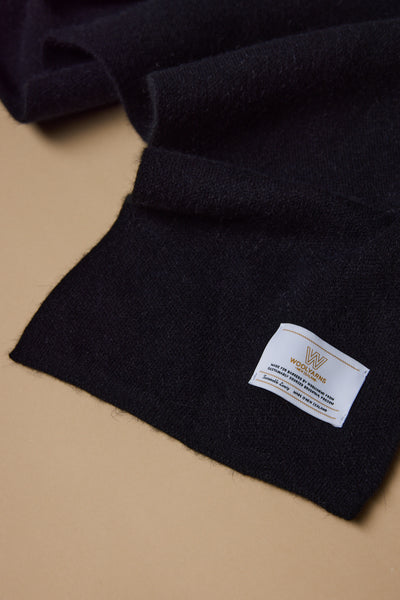 A close-up of the NZ Made Perino Scarf by Woolyarns Perino, in black wool and a luxurious merino possum silk blend, on a beige surface with a white label and gold WOOLMARK logo—crafted in New Zealand.