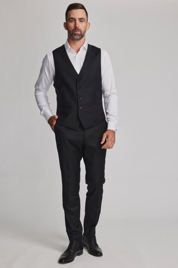 A man with short hair and a beard stands against a light gray background, wearing a white dress shirt, black dress pants, black dress shoes, and the Barkers Mayfair ZQrx Waistcoat with one hand in his pocket for a refined look.