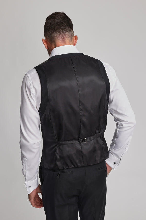 A man is seen from behind wearing the Barkers Mayfair ZQrx Waistcoat in black over a white shirt and black trousers. His right hand is on his hip, showing the vest’s adjustable strap. The background is plain and light-colored.