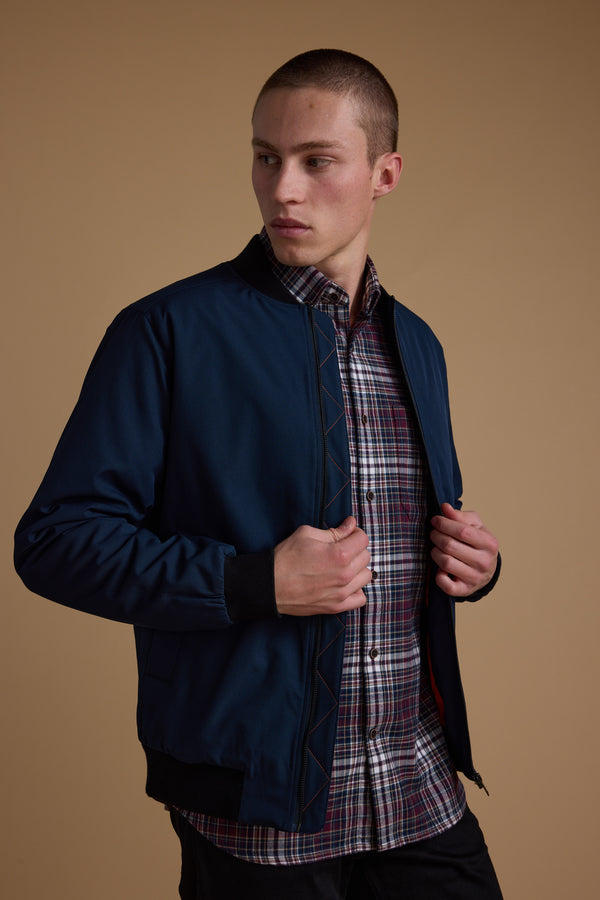 A young man models the Barkers Dunhill Bomber in navy blue recycled polyester over a plaid shirt, standing against a beige background and looking left while holding the jacket open.