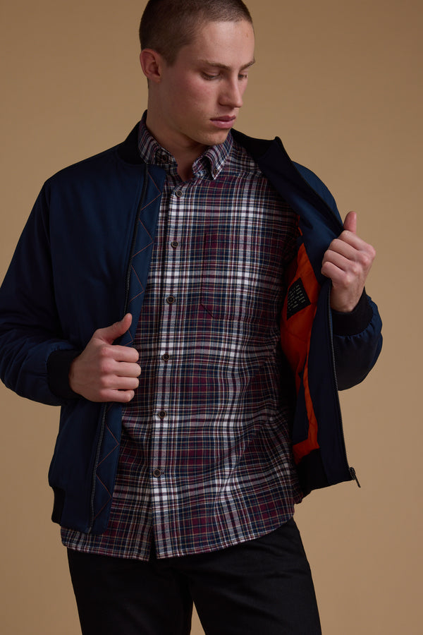 Someone wearing the Barkers Dunhill Bomber, a navy recycled polyester jacket with an orange inner lining, opens it over a plaid shirt against a neutral beige background.