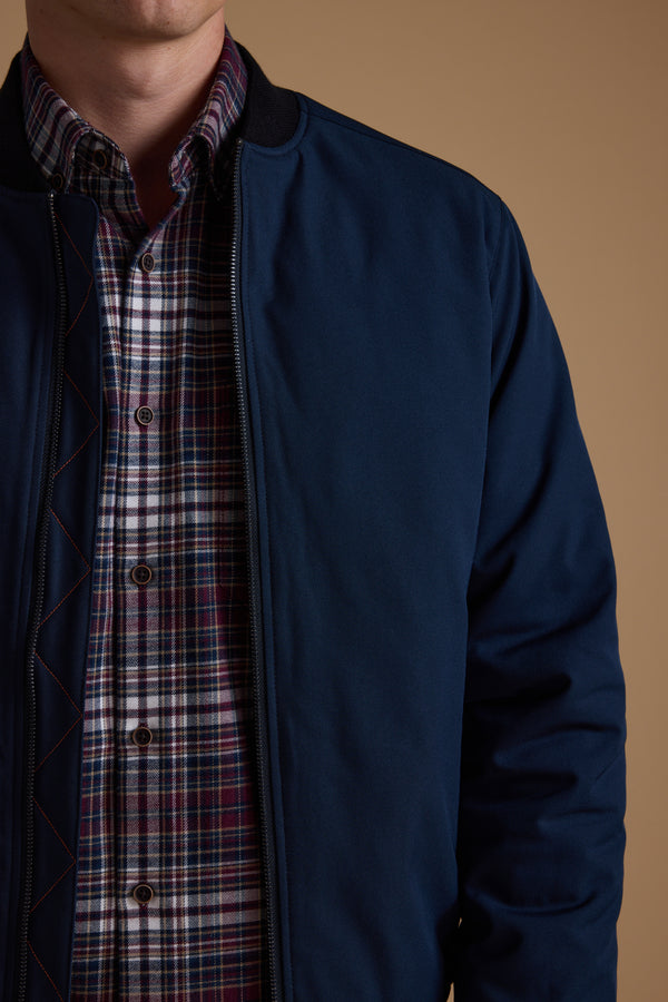 A person wears the Barkers Dunhill Bomber, a blue recycled polyester jacket, over a red and white plaid shirt, shown from shoulders to waist against a plain beige background.