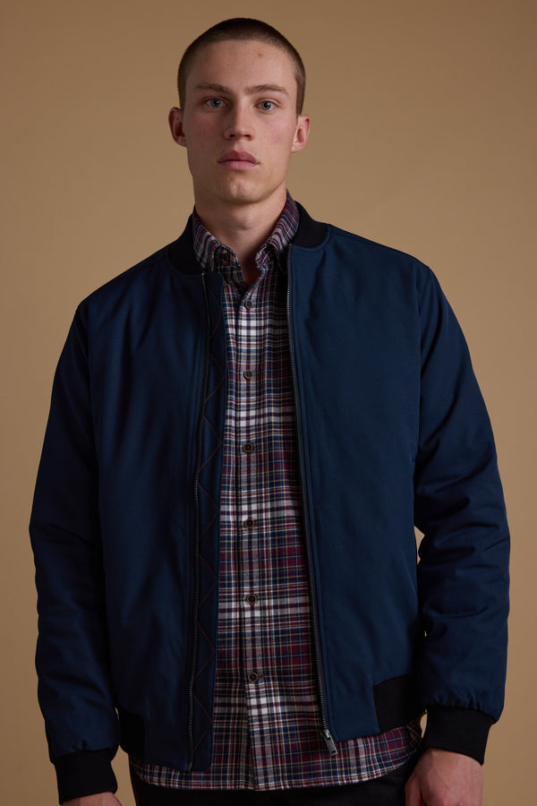 A young man with short hair models the Barkers Dunhill Bomber, a navy blue water-resistant jacket, over a red and white plaid shirt against a beige background, facing the camera with a neutral expression.