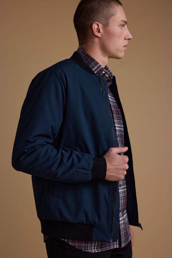 A person in profile wears a navy blue Barkers Dunhill Bomber over a plaid shirt, standing against a plain beige background.
