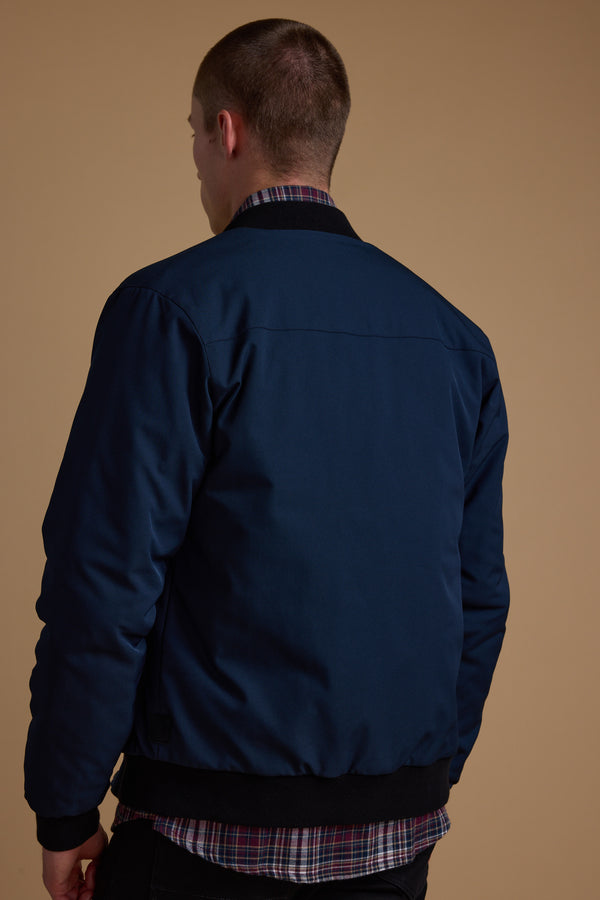 A person with short hair faces away from the camera, wearing a navy blue Barkers Dunhill Bomber over a red and blue plaid shirt, standing against a plain beige background.