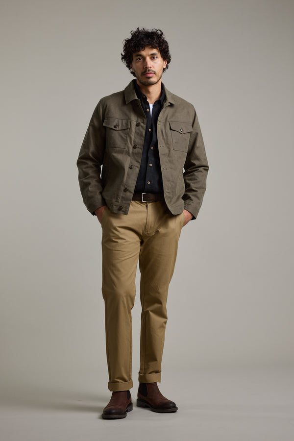 A man with curly dark hair stands against a plain background, wearing the Barkers Benson Waxed Cotton Jacket in khaki over a dark shirt, beige pants, and brown shoes. His hands are in his pockets as he faces the camera.
