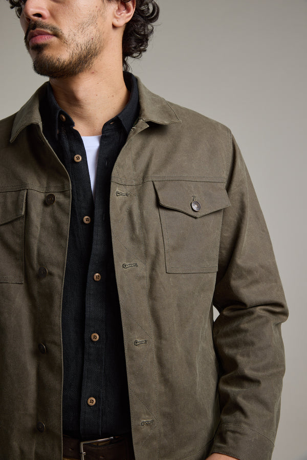 A man with short curly hair wears a Barkers Benson Waxed Cotton Jacket in olive green over a black button-up and white undershirt. Shown from shoulders to mid-thigh, he faces slightly sideways against a plain background.