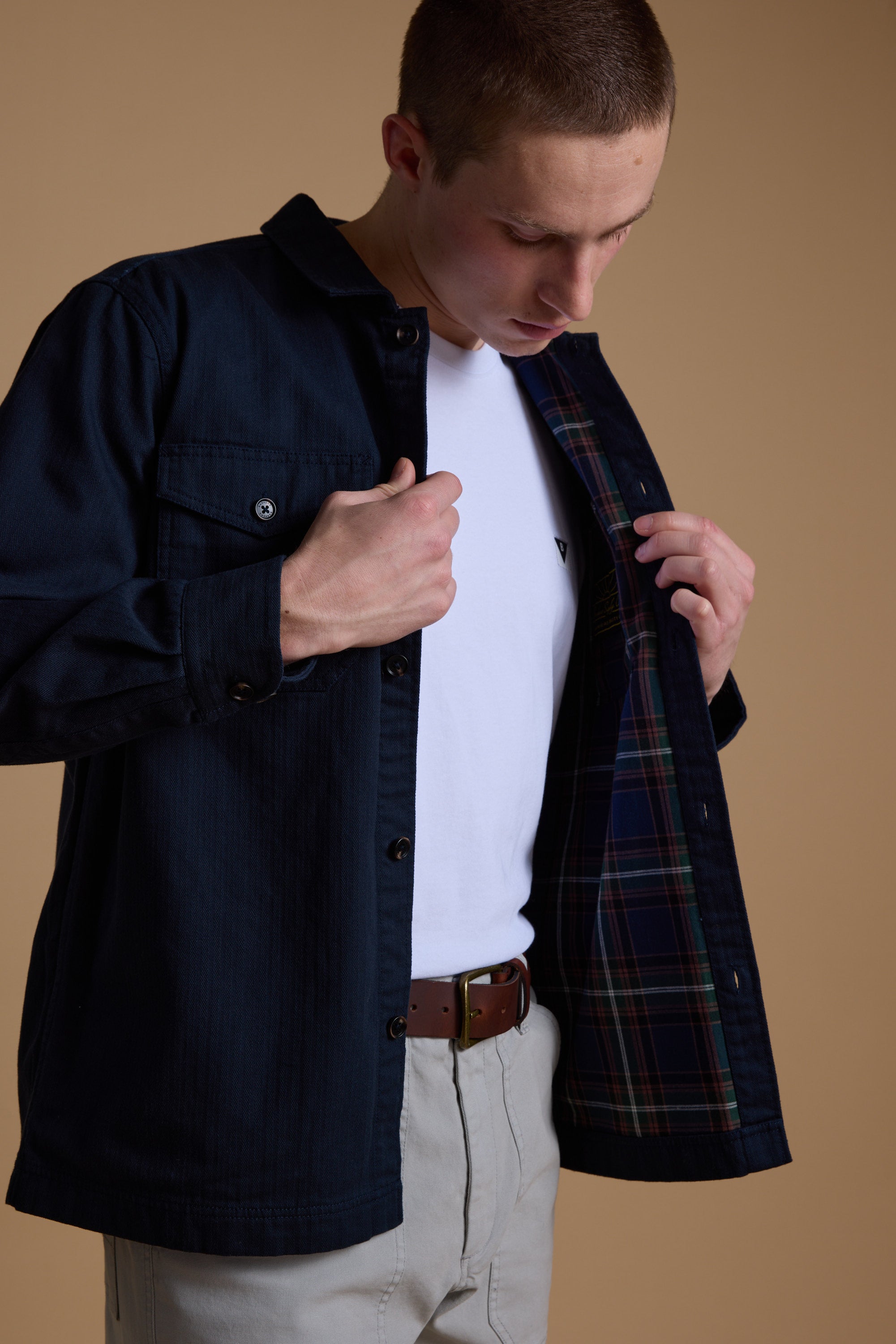 A person wearing the Barkers Gifford Herringbone Jacket over a white t-shirt and beige pants looks down, holding the jacket open to show its flannel lining against a solid tan background.