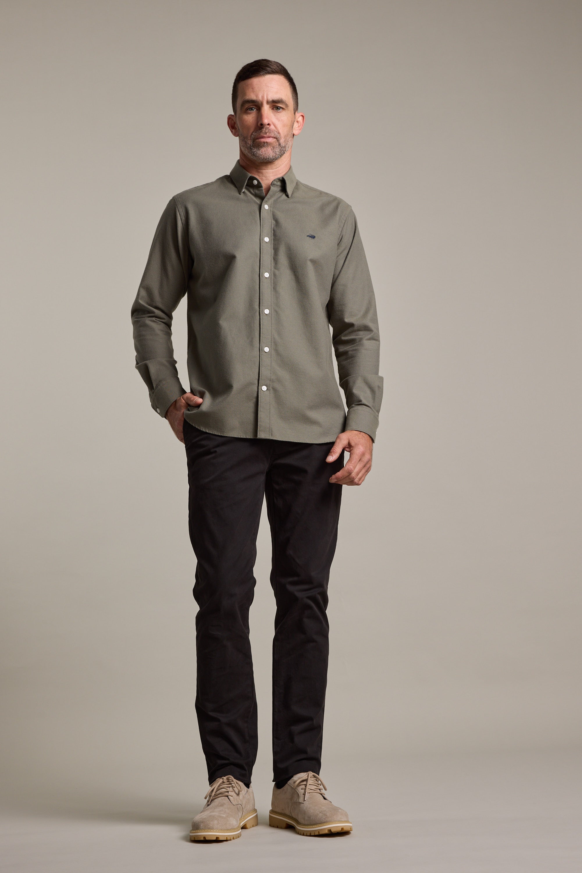 A man stands facing forward in an olive-green button-up shirt, Barkers Carnaby Chino trousers in black, and beige shoes, with one hand in his pocket against a plain light background.